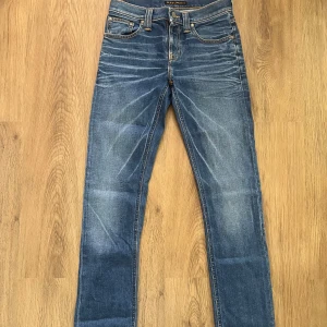 Nudie Jeans  - Snygga blåa jeans från Nudie Jeans med klassiska orange sömmar och ikonisk brodyr på bakfickorna. Jeansen har normal midja och är tillverkade i mjukt denimtyg. Perfekta för dig som gillar en stilren look.                                                Storleken är W26 L34 och jeansen är endast använda några gånger. Passformen är Slim Fit.