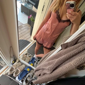 Rosa linnetopp med spetsdetaljer - Söt rosa linnetopp och shorts med svarta spetsdetaljer längs urringningen och tunna axelband. Toppens material känns lätt och luftigt, perfekt för varma dagar eller som en snygg layering-piece. Den har en feminin v-ringning och en avslappnad passform.