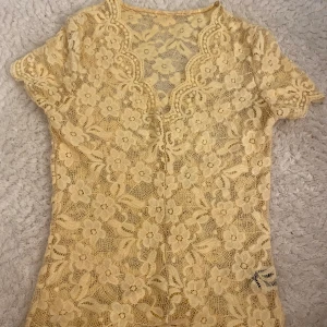 Gul spetsblus med blommönster - Supersöt gul blus i spets med blommigt mönster och korta ärmar. Blusen har v-ringning och är genombruten med fina detaljer i spetsen. Perfekt för dig som vill sticka ut med en romantisk vibe.