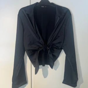 Svart satin omlottblus från Bik Bok - Stilren svart omlottblus från Bik Bok i glansigt satintyg. Blusen har långa ärmar, klassisk krage och knyt framtill för en snygg och trendig look. Perfekt att styla med jeans eller kjol för en chic vibe. Justerbar storlek. Passar s-2xl. Nypris 399kr aldrig använd