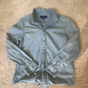 Ljusblå skjorta från Ralph Lauren - Snygg ljusblå skjorta från Ralph Lauren i bomull. Storlek 14 sitter som S. Klassisk modell med knappar hela vägen, lång ärm och broderad logga på bröstet. Perfekt för en clean och avslappnad look.