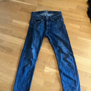 Blå jeans från Dressmann Regular Fit - Snygga blå jeans från Dressmann i modellen Regular Fit. Klassisk femficksdesign med dragkedjegylf och kontrastsömmar. Jeansen har en rak passform och är tillverkade i slitstarkt denimtyg som passar till det mesta.