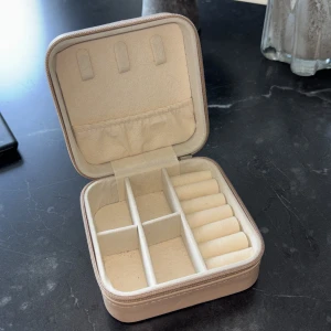 Beige smyckesväska i skinnimitation - Snygg och praktisk beige smyckesväska i fyrkantig form med dragkedja runtom. Insidan har flera fack och rullar för ringar, perfekt för att hålla koll på dina smycken när du är på språng. Stilren design som passar i väskan eller på resan.