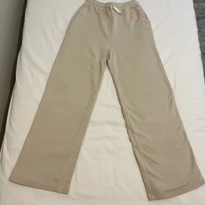 Beiga mjukisbyxor med LA-detalj - Snygga beiga mjukisbyxor med breda ben och broderad LA-logga i rosa framtill. Byxorna har resår i midjan med snörning för extra komfort. Perfekta för en chill och avslappnad stil.