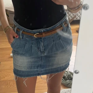 Blå jeanskjol med fransig kant - Säljer en snygg blå jeanskjol med fransig nederkant och coola slitningar. Kjolen har fickor fram och bak samt hällor för bälte, och kommer med ett brunt bälte. Perfekt för dig som gillar en avslappnad och trendig stil.