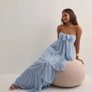Ljusblå långklänning med volanger storlek S - Superfin ljusblå långklänning med flera lager volanger och knytning vid bysten. Klänningen är axelbandslös framtill och har halterneck med rosett i nacken. Luftigt material som ger en flowy känsla, perfekt för varma dagar.