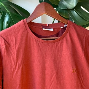 Röd t-shirt med broderad detalj - Snygg röd t-shirt i bomull med korta ärmar och rund halsringning. På bröstet finns en liten broderad detalj i form av två romerska siffror. Enkel och stilren modell som passar perfekt till jeans eller shorts.