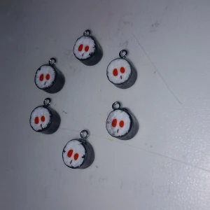 Sushi-inspirerade berlocker - Sex stycken handgjorda berlocker som ser ut som små sushibitar med svart, vitt och rött. Perfekta att använda som hängen till halsband eller armband för en unik och lekfull stil.