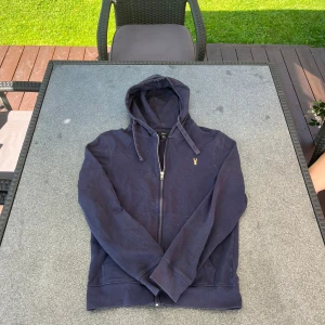 Mörkblå hoodie från Lyle & Scott - Mörkblå hoodie från Lyle & Scott med dragkedja och huva. Klassisk gul logga broderad på bröstet, känguruficka framtill och ribbade muddar. Perfekt för chill dagar och enkel att matcha med jeans eller joggers.