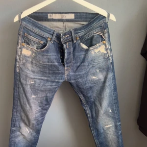 Blå slitna jeans från Replay - Snygga blå jeans från Replay med slitna detaljer och klassisk femficksmodell. Jeansen har ljus tvätt, gylf med knappar och broderade bakfickor. Perfekta för en avslappnad streetstil och passar dig som gillar en trendig look.