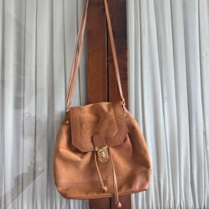 Brun mocka axelväska med snörning - Snygg axelväska i brun mocka med detaljer i skinn. Väskan har snörning och ett lock med spänne i metall. Perfekt storlek för att få plats med allt du behöver och har en avslappnad men stilren look.