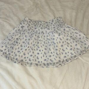 Vit blommig kortkjol från Gina Tricot - Supersöt vit kortkjol från Gina Tricot med blått blommönster. Kjolen har en flowy passform och resår i midjan för extra komfort. Perfekt för varma dagar och ger en fräsch och somrig vibe. Materialet känns lätt och luftigt.