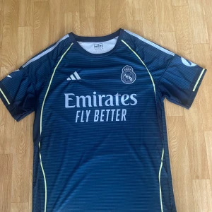 Real Madrid  S - Blå Real Madrid fotbollströja från Adidas med Mbappé och nummer 9 på ryggen. Tröjan har tunna horisontella ränder, gula detaljer längs sidorna och vita loggor. Tillverkad i lätt och ventilerande polyester, perfekt för träning eller match.