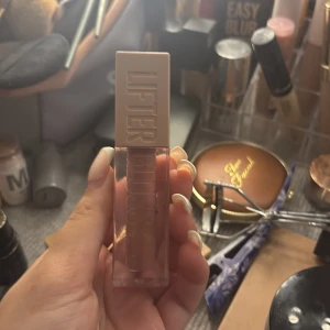 Maybelline Lifter Gloss nude - Superfin Lifter Gloss från Maybelline i en nude/ljusrosa nyans. Kommer i en stilren fyrkantig förpackning med beige lock och stor applikator. Perfekt för glansiga läppar med en hint av färg. 