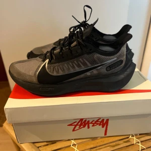 Nike Zoom svart/grå sneakers - Nike Zoom sneakers i svart och grått med genomskinlig mesh, stora svarta swoosh-loggor och reflekterande detaljer. Skorna har snörning och en chunky sula för extra komfort. Perfekta för dig som gillar sportig och modern stil.