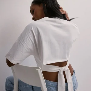 Back tie Nelly tröja - Snygg vit croppad topp i bomull med korta ärmar och cool knytning i ryggen. Enkel och stilren design som passar perfekt till jeans eller kjol. Toppen har en rak passform framtill och öppen rygg med breda knytband för en trendig look.