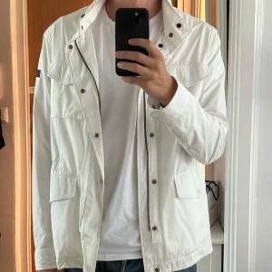 Vit lättviktsjacka från Hetregó - Snygg vit vindjacka med utfällbar luva från Hetregó. Jackan är i övrigt i bra skick, men är smutsig på sina ställen, t.ex vid kragen och bröstfickorna (båda går bort vid tvätt). Jackan är köpt på United fashion i Göteborg för ca 3500kr. Skriv vid funderingar!