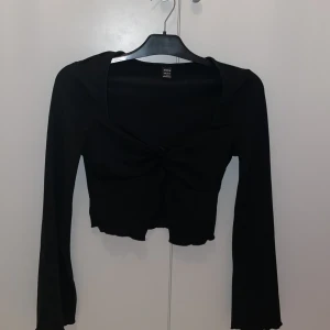 Svart croppad topp från SHEIN - Liknar ginas topp. Svart långärmad croppad topp från SHEIN med twistad detalj framtill. Toppen har en lätt v-ringad halsringning och vågig kant nertill. 