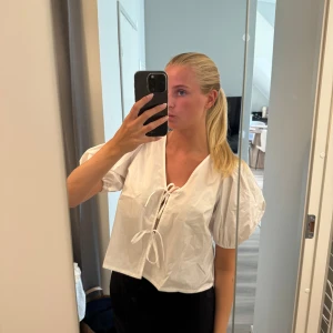 Vit blus med puffärm och knyt - Säljer en vit blus med korta puffärmar och v-ringning. Blusen har två knyt framtill som ger en söt och trendig look. Modellen är croppad och har en luftig passform, perfekt för varmare dagar.