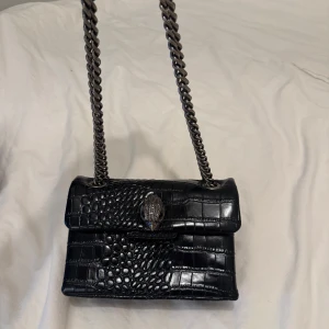 Svart axelväska från Kurt Geiger - Säljer en svart axelväska i äkta skinn från Kurt Geiger med croco-mönster och en chunky silverfärgad kedja. Väskan har ett snyggt spänne framtill i form av ett dekorerat huvud och magnetlås. Perfekt storlek för det viktigaste och ger en lyxig vibe.