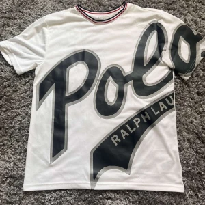 Vit Polo Ralph Lauren t-shirt med tryck - Vit t-shirt från Polo Ralph Lauren med stort svart logotyp-tryck över hela framsidan. T-shirten har korta ärmar, rund hals med röd och marinblå randig kant och är tillverkad i mjuk polyester.