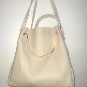 Set med tre beige väskor - Säljer ett set med tre matchande beige väskor i syntetmaterial. En större axelväska med handtag och metall-detaljer, en mindre väska med justerbar rem och tofs, samt en kuvertväska med kedja och metallplatta framtill. Perfekt för dig som vill ha stilrena och enkla accessoarer.