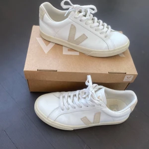 Vita Veja sneakers med beige detaljer - Snygga vita sneakers från Veja med beige mockadetaljer på sidorna och hälen. Klassisk låg modell med snörning och platt sula. Perfekta för en clean och stilren look. Ikoniskt V på sidan och diskret branding på sulan.