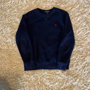 Mörkblå sweatshirt från Polo Ralph Lauren - Klassisk mörkblå sweatshirt från Polo Ralph Lauren med rund hals och röd broderad logga på bröstet. Tröjan har långa ärmar och ribbade muddar vid ärmslut och nederkant. Perfekt för en stilren och avslappnad look.