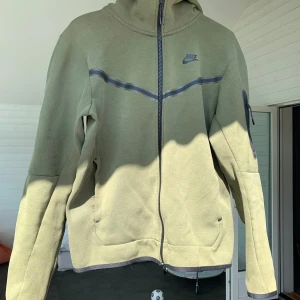 Nike tech fleece i grönt - Nike techfleece i olivgrönt med hel dragkedja och huva. Jackan har en mörkgrå detalj över bröstet, fickor i sidorna och en extra ficka med dragkedja på ärmen. 