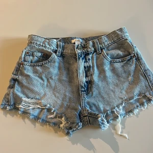 Blå jeansshorts från Gina Tricot - Snygga ljusblå jeansshorts från Gina Tricot med fransig och sliten look. Klassisk femficksmodell med bälteshällor och råa kanter nertill. Perfekta för en avslappnad och trendig stil i sommar.strl 152