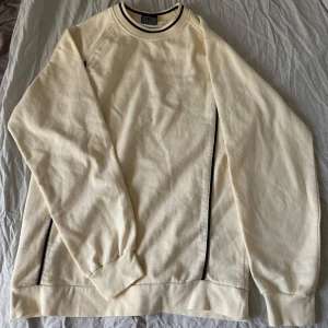 Beige sweatshirt från New Wave S - En stilren beige sweatshirt från New Wave i storlek small. Tröjan har svarta ränder vid kragen och längs sidorna, rund hals och långa ärmar. Materialet är en skön mix av 70% bomull och 30% polyester. Perfekt för en clean och avslappnad look.