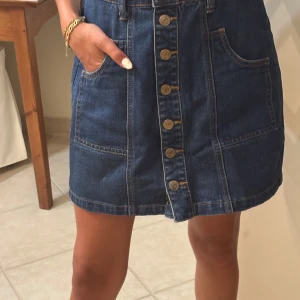 Jeanskjol med knappar från Stradivarius - Snygg mörkblå jeanskjol från Stradivarius med raka linjer, stora fickor framtill och dekorativa knappar hela vägen ner. Kjolen är kort och har klassiska kontrastsömmar som ger en cool vintagekänsla. Perfekt för dig som gillar en avslappnad men trendig stil.