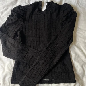 Svart spetsblus med puffärm - Snygg svart blus i spets med lång ärm och diskreta puffärmar. Blusen har ett rutigt mönster i spetsen och stängs med knapp i nacken. 