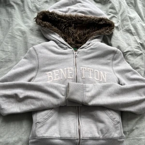 Grå hoodiejacka från Benetton - Mysig grå hoodiejacka från Benetton med dragkedja och broderad logga framtill. Jackan har huva fodrad med mjuk pälsimitation och ribbade muddar vid ärmslut och nederkant. Perfekt för kyliga dagar.
