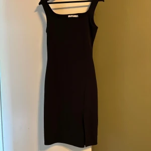 Svart kort klänning från NA-KD - En stilren svart kort klänning från NA-KD med breda axelband och fyrkantig ringning. Klänningen har en smickrande slim passform och en liten slits framtill för extra edge. Perfekt för dig som gillar minimalistisk och tidlös stil.