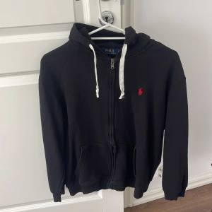 Polo hoodie - Svart hoodie från Polo Ralph Lauren med vit snörning i huvan och röd broderad logga på bröstet. Tröjan har dragkedja framtill, känguruficka och långa ärmar. Perfekt för en chill och stilren look.