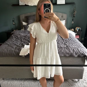 Vit spetsklänning från ONLY - Superfin vit kortklänning från ONLY med v-ringning, volangärmar och spetsdetaljer. Klänningen har knappar framtill och ett luftigt, romantiskt fall. Perfekt för sommarens alla tillfällen och ger en fräsch och söt look.