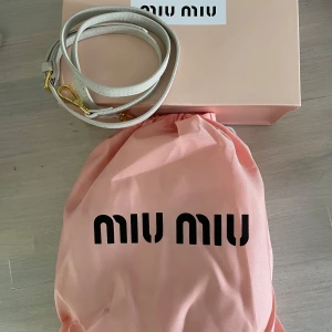 Vit mocka handväska från Miu Miu - Supersnygg vit handväska från Miu Miu i mjuk mocka med guldiga metalldetaljer och dragkedja. Väskan har två stadiga handtag och präglad logga framtill. Perfekt storlek för det viktigaste och ger en clean, lyxig vibe. Dustbag och axelband följer med.