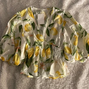 Vit blus med citronmönster och puffärm - Superfin vit blus från H&M med citronmönster i gult och grönt. Blusen har v-ringning, smockad midja och stora puffärmar som ger en riktigt somrig vibe. Perfekt för dig som vill sticka ut med färg och mönster! Helt ny och oanvänd med prislappen kvar. Slutsåld på hemsidan