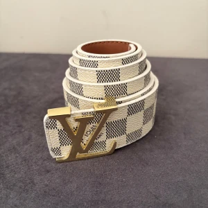 Rutig Louis Vuitton bälte med LV-spänne - Snyggt bälte från Louis Vuitton med klassiskt rutigt mönster i vitt och grått. Bältet har ett stort guldfärgat LV-spänne och är gjort i skinn med ljusbrun insida. Perfekt accessoar för att lyfta din outfit med en lyxig touch.