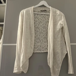 Vit kofta med spetsrygg - Säljer en vit kofta från Lindex med långärmad design och öppen front. Baksidan har ett snyggt spetsmönster som ger en extra touch. Perfekt att slänga över en topp för en chill och stilren look.