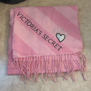 Rosa randig halsduk Victoria's Secret - Superfin rosa halsduk från Victoria's Secret med breda diagonala ränder i olika rosa nyanser. Halsduken har fransar i ändarna och en glittrig hjärtapplikation med strasstenar. Perfekt accessoar för att lyfta din outfit.