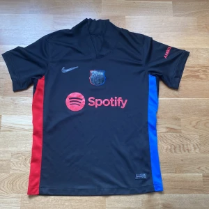 FC Barcelona svart Nike fotbollströja - Snygg svart FC Barcelona fotbollströja från Nike med röda och blå sidopaneler. Tröjan har klubbmärke på bröstet, Spotify-logga framtill och UNHCR-tryck på ryggen. Kortärmad modell i lätt och ventilerande material, perfekt för match eller träning.