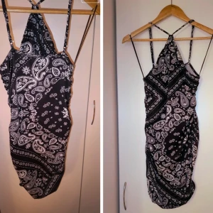 Svart kort klänning med paisleymönster - Säljer en svart kort klänning med vitt paisley- och bandanamönster. Klänningen har smala axelband som korsas i ryggen och en halterneck-liknande överdel. Sitter tight och har en snygg, figurnära passform. Perfekt för dig som gillar unika mönster och vill sticka ut.