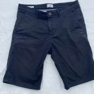 Svarta shorts från Jack & Jones - Klassiska svarta shorts från Jack & Jones med normal passform. De har två sidofickor, två bakfickor och stängs med knapp och dragkedja. Perfekta för sommaren och tillverkade i mjuk bomull för en skön känsla.