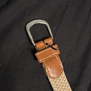 Beige flätat bälte med skinn-detaljer - Snyggt beige flätat bälte med bruna skinn-detaljer och silvrigt spänne. Perfekt accessoar för att lyfta din outfit med en stilren och klassisk touch. Bältet har en vävd struktur och passar till både jeans och chinos.