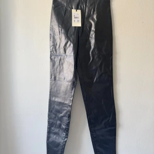 Svarta byxor i läder imitation  - Oanvända, prislapp kvar. Går att öppna nere vid benen för att få en ”slits look”.  Ca 100cm i längd Från sida till sida vid byxöppning ca 33cm.