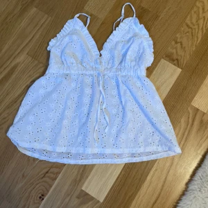 Vit broderad linnetopp från new yorker  - Supersöt vit linnetopp från new yorker med broderade blommor och volangdetaljer vid axelbanden. Toppen har v-ringning, smala axelband och knyt i midjan för en extra snygg passform. Perfekt till sommaren och riktigt fräsch till shorts eller kjol.
