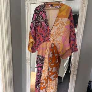 Vacker sommarklänning från Zara - Säljer en jättefin klänning från Zara med färgstarkt blommigt och paisleymönster i orange, rosa, lila och beige. Klänningen har v-ringning med pärldetaljer. Smickrande passform. Perfekt för dig som vill sticka ut med en bohemisk vibe. Oanvänd. 