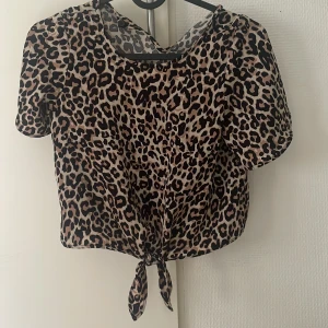 Leopardmönstrad blus från Lindex Kids - Snygg blus från Lindex Kids i leopardmönster med korta ärmar och knyt framtill. Toppen är i beige och svart, har rund halsringning och är tillverkad i ett lätt, syntetiskt material. Perfekt för dig som vill sticka ut med ett djurmönster.